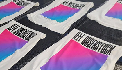 White t-shirts on a conveyor dryer belt that have a pink to purple to cyan blue gradient with the text Jeff Rosenstock screen printed onto it.