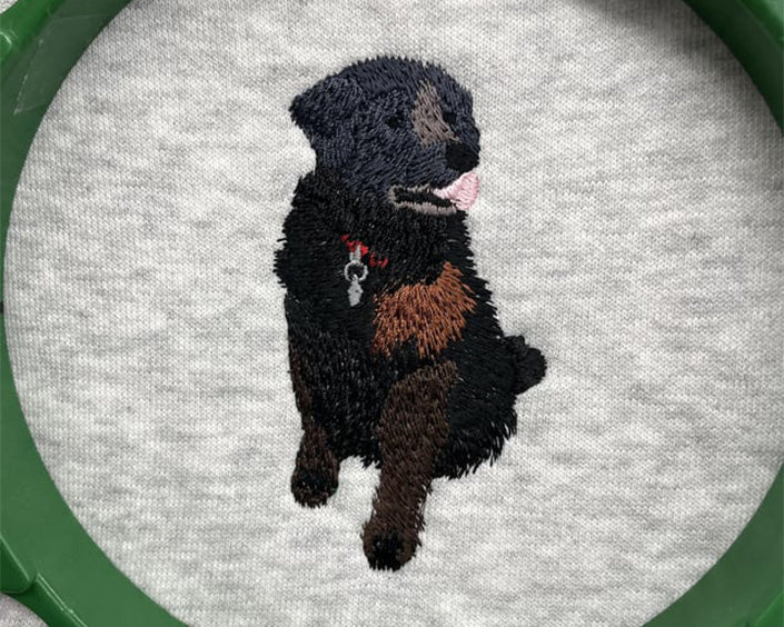 Production of Sampson sewn out onto a sweatshirt. The dog has his tongue sticking out with a nice bright red collar and silver tag against his black fur. 